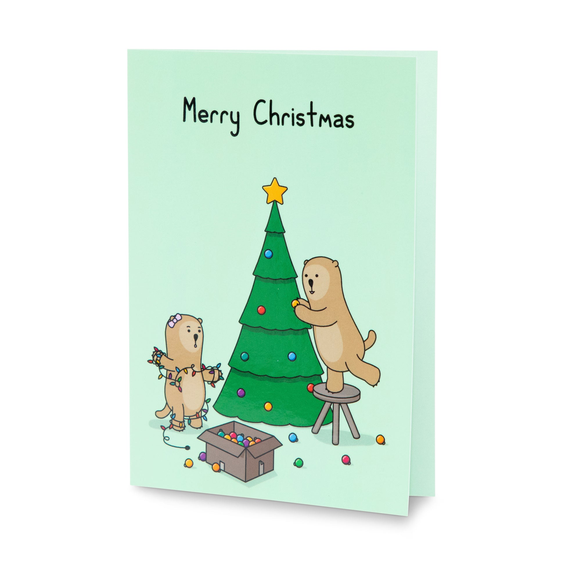 A green Christmas greeting card with a cheerful illustration of two otters decorating a Christmas tree. One is reaching to put decorations on the tree, the other is tangled in Christmas lights. The text on the card reads 'Merry Christmas'.