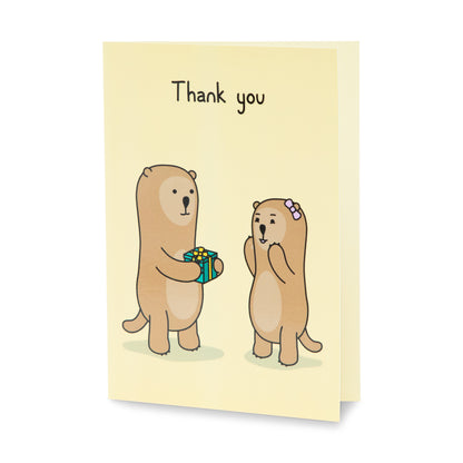 A yellow thank you greeting card with a cheerful illustration of one otter handing a green present to an exited otter. The text on the card reads 'Thank you'.