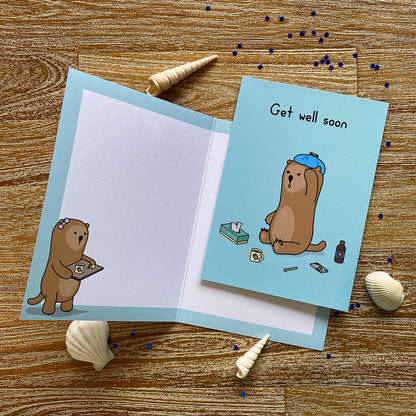 A blue get well soon greeting card with an image of an otter sitting on the ground with an icebag on his head. He is surrounded by various items, a tissue box, thermometer, a box of pills and a bottle of medicine. The text on the card reads 'Get well soon'.