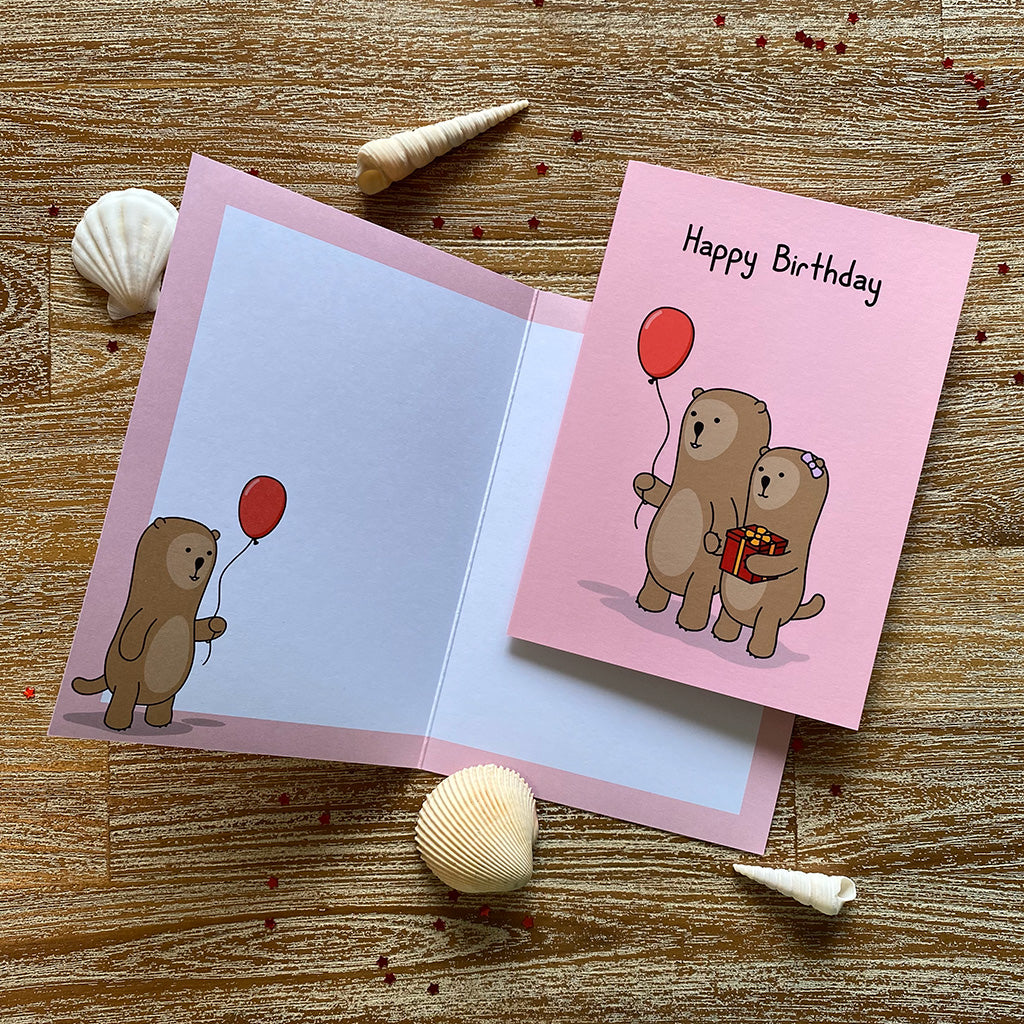 A red birthday greeting card with a cheerful illustration of otters going to a party one holding a balloon, the other holding a present. The text on the card reads 'Happy Birthday!'.