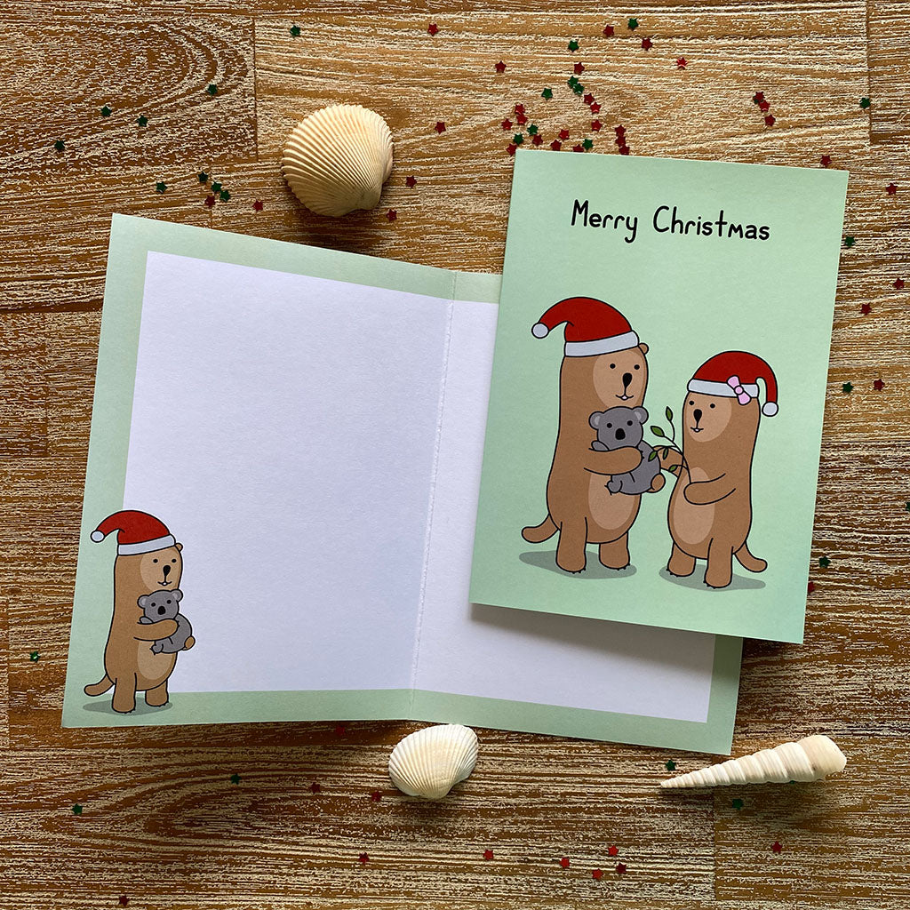 A green Christmas greeting card with a cheerful illustration of two otters in Santa hats, one is holding a koala, the other is feeding the koala leaves. The text on the card reads 'Merry Christmas'.