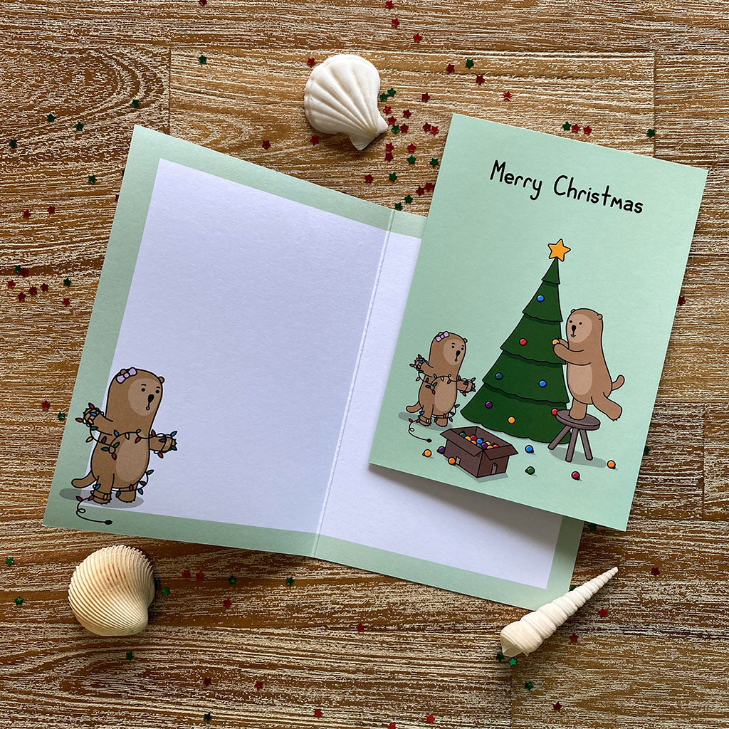 A green Christmas greeting card with a cheerful illustration of two otters decorating a Christmas tree. One is reaching to put decorations on the tree, the other is tangled in Christmas lights. The text on the card reads 'Merry Christmas'.