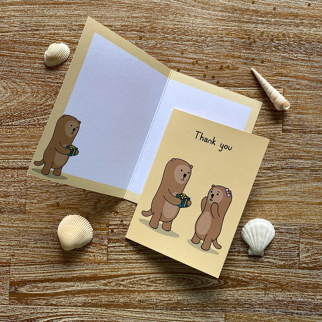 A yellow thank you greeting card with a cheerful illustration of one otter handing a green present to an exited otter. The text on the card reads 'Thank you'.